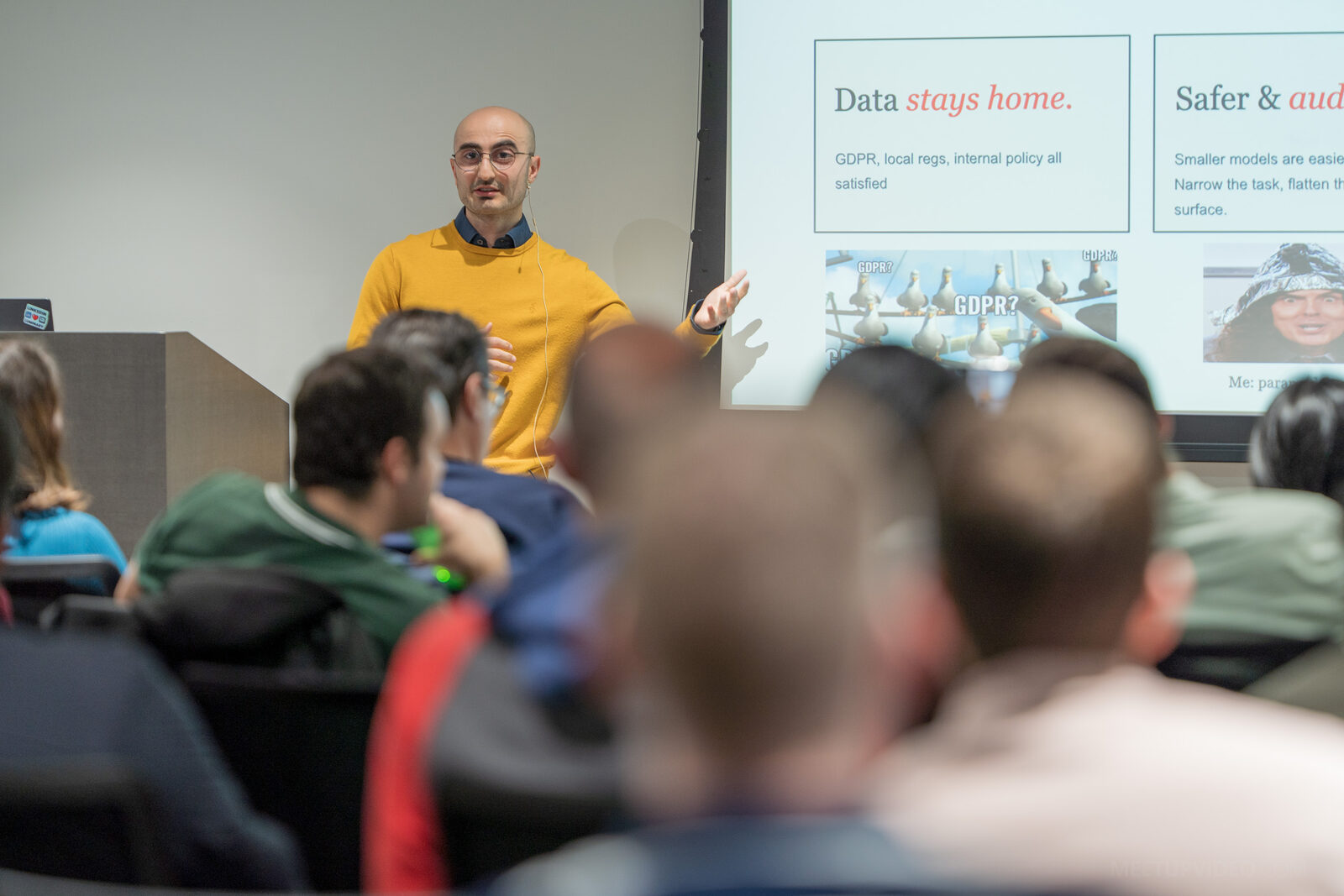 A speaker in a yellow jumper gesturing towards a slide reading “Data stays home” — GDPR, local regs and internal policy all satisfied — alongside a panel on smaller models being safer and auditable.
