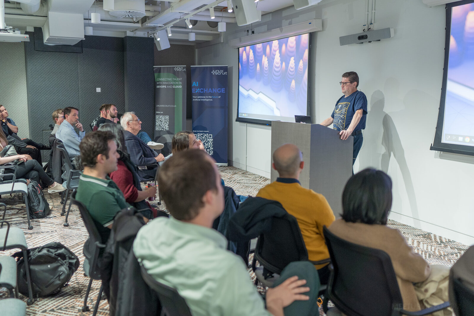 A speaker at the lectern mid-talk, the audience in the foreground and AI Exchange and Linux Recruit pull-up banners standing beside the screen.