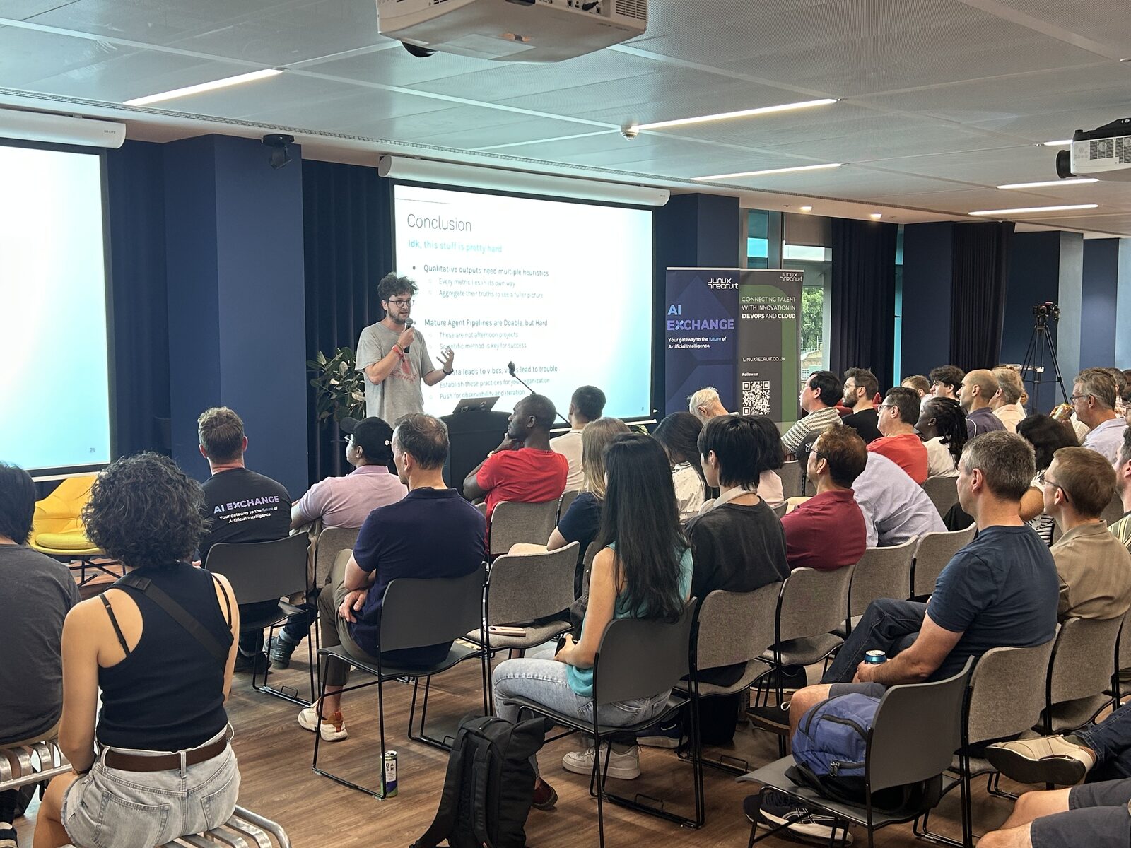 A full room at AI Exchange @ Tessl, the audience seated in rows facing a speaker and a projected slide.